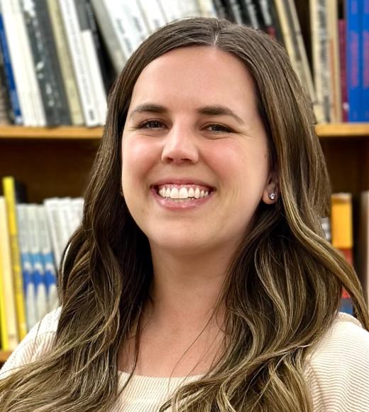 Professional photo of a smiling young woman with brown hair. There are books on a shelf in the background