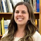 Professional photo of a smiling young woman with brown hair. There are books on a shelf in the background